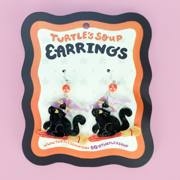 A pair of black cat witch earrings with purple hats and orange shoes, displayed on a decorative black, orange, and white backing card that reads 'TURTLE'S SOUP EARRINGS' against a solid pink background.