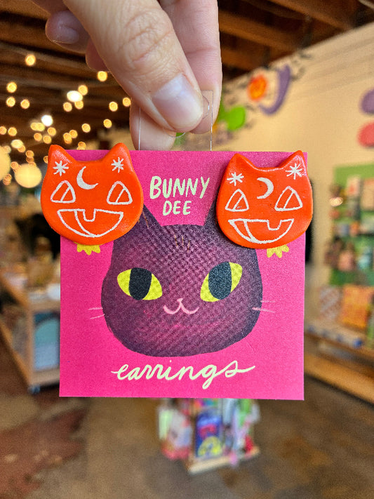 A hand holds a pink card featuring a black cat face with yellow eyes and orange pumpkin-eared earrings attached. The card reads 'BUNNY DEE earrings' and is photographed in a softly blurred store setting with warm lights in the background.