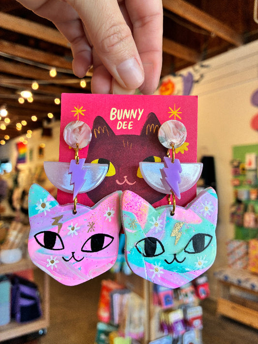 A hand holds a pair of colorful acrylic cat-ear earrings in front of a pink backing card labeled 'BUNNY DEE' inside a shop. The earrings have pastel pink and mint green cat heads with white daisy patterns, purple lightning-bolt shapes, and iridescent round studs.