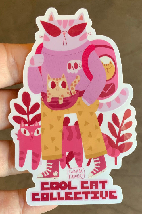 A hand holding a die-cut sticker featuring a pink and orange cat character with the text 'COOL CAT COLLECTIVE' at the bottom, against a brown concrete floor background.