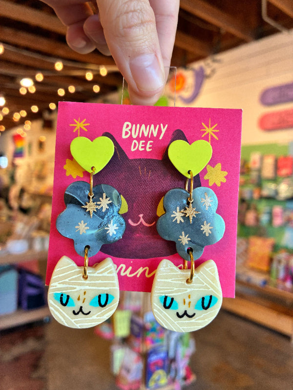 A hand holds a pink card displaying a pair of colorful cat-themed drop earrings in a shop setting. The earrings feature lime green heart studs, blue flower charms with gold star details, and cream-colored mummy-wrap cat pendants with blue eyes.