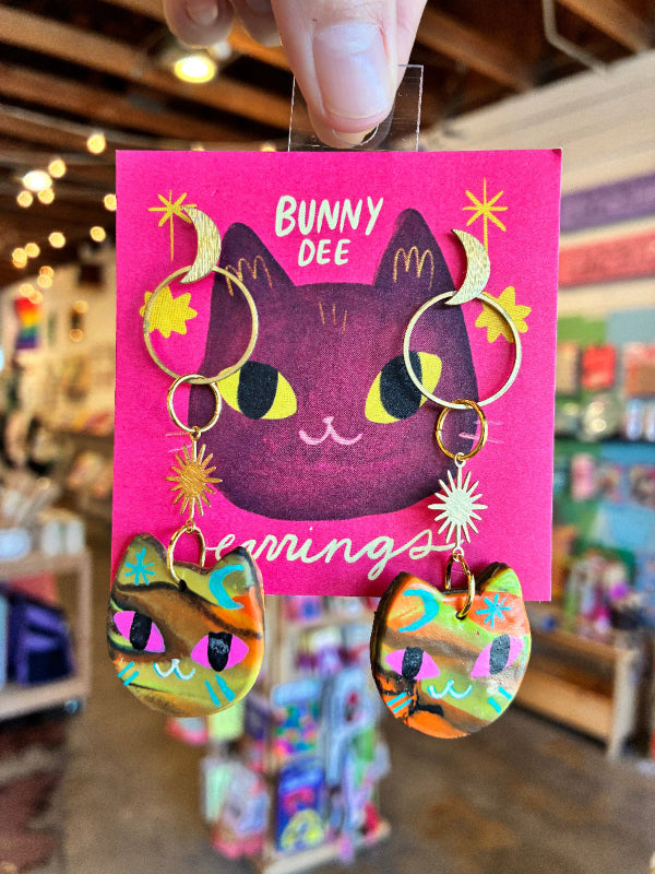 A hand holds a pink display card featuring a pair of colorful, hand-painted bunny-shaped earrings with gold-tone rings and crescent moon charms. The background is a blurred shop interior.