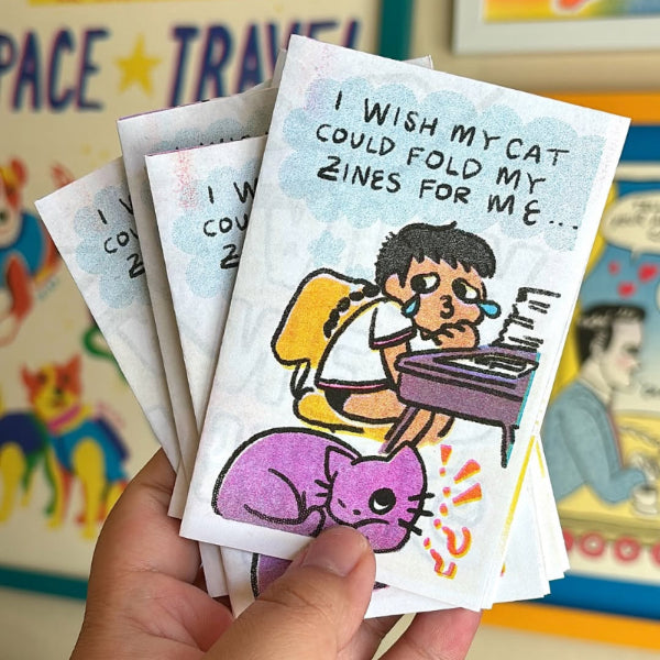 Hand holding a set of zine pages with a cartoon character and cat, against a colorful background.